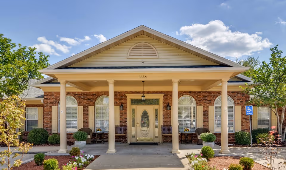 Front exterior view of a single-story brick building with a covered porch supported by four columns. The building has a central door with decorative glass and four arched windows with shutters. There are chairs and small tables on the porch, landscaping with bushes and flowers, and a handicapped parking sign visible on the right side.