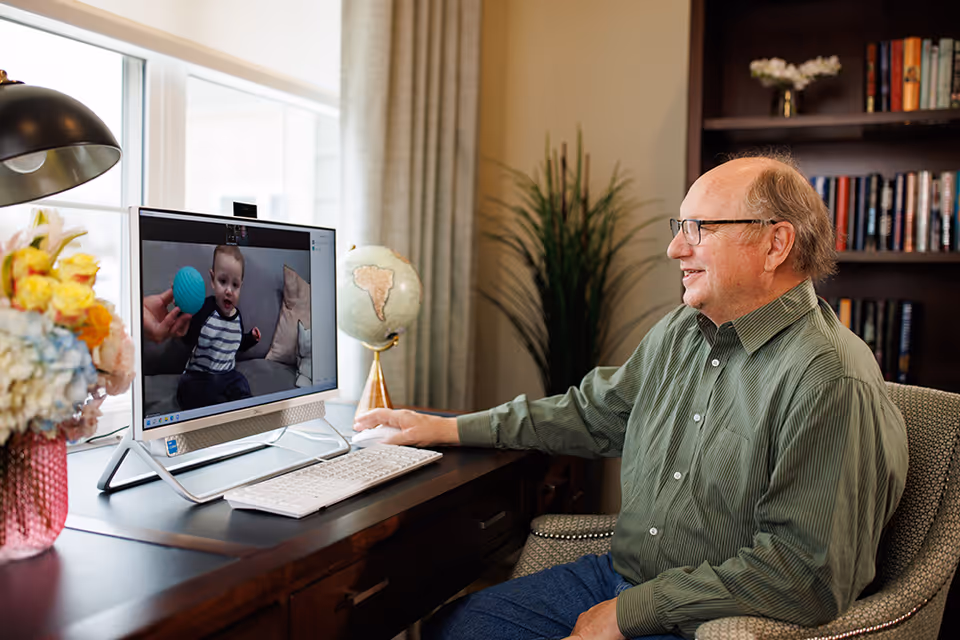 An older man sits at a desk smiling while video-calling a baby shown on a desktop computer in a cozy, book-lined room.