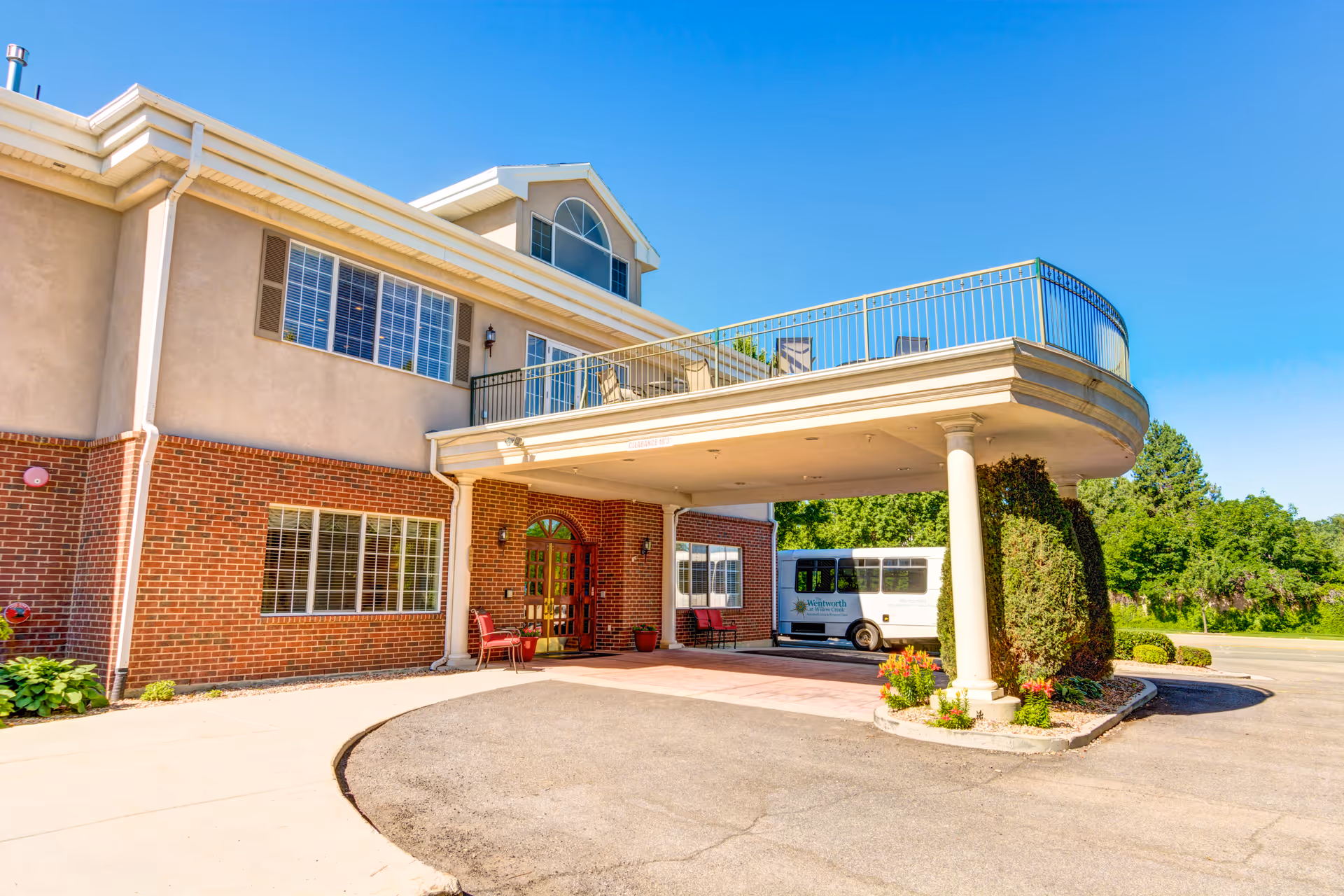 Front entrance of a senior living facility with a covered porte-cochere, balcony above, and a shuttle van parked nearby.