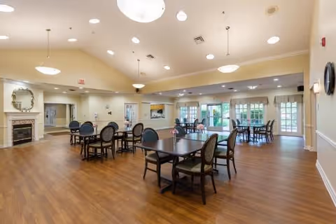 Spacious dining area with multiple dark wood tables and chairs arranged on a wood floor. The room has cream-colored walls, large windows with curtains, several ceiling lights, and a fireplace with a decorative mirror above it. The space is well-lit with natural and artificial light.