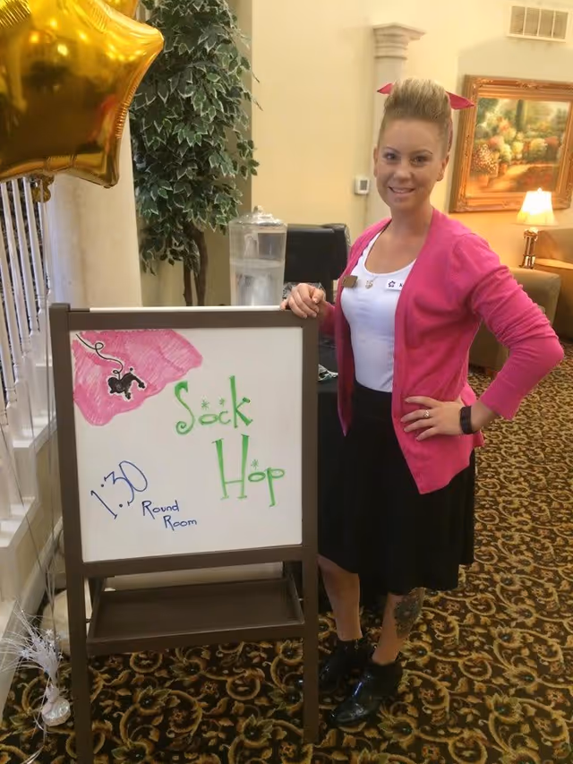 A woman dressed in a pink cardigan, white top, and black skirt stands next to a whiteboard sign that reads 'Sock Hop 1:30 Round Room' with a drawing of a pink sock and a black poodle. The setting appears to be an indoor common area with patterned carpet, a plant, and framed artwork on the wall.