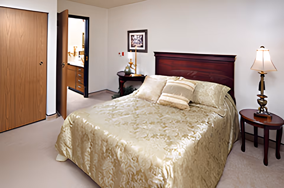 A furnished bedroom with a bed covered in a gold bedspread, bedside tables with lamps, and a doorway leading to a bathroom.