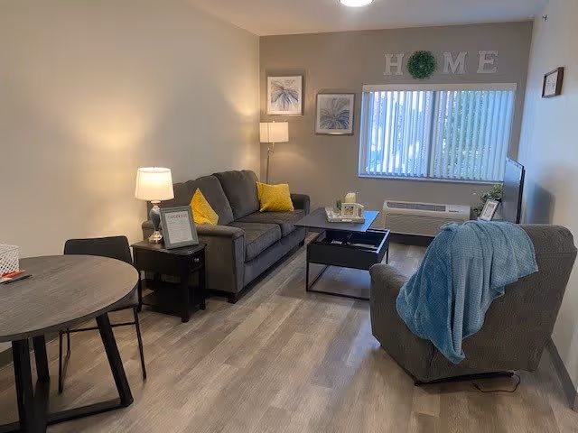 A cozy living room with a gray sofa adorned with yellow pillows, a matching armchair draped with a blue blanket, a coffee table, and a side table with a lamp. There is a round dining table with chairs on the left side. The wall has two framed pictures and a decorative 'HOME' sign above a window with vertical blinds. The floor is light wood, and the room is softly lit by lamps.
