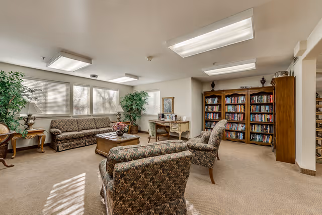 A cozy living room area with patterned upholstered chairs and a sofa, a wooden coffee table with a flower arrangement, a desk with a chair near the window, and a large bookshelf filled with books. The room is well-lit with natural light coming through multiple windows and ceiling lights.