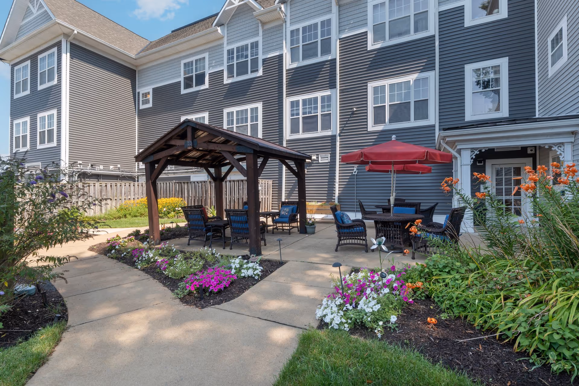 Outdoor patio area at Sunrise of Springfield featuring a wooden pergola with seating underneath, a round table with chairs and a red umbrella, surrounded by flower beds with colorful flowers and greenery, adjacent to a multi-story gray building with white-trimmed windows.