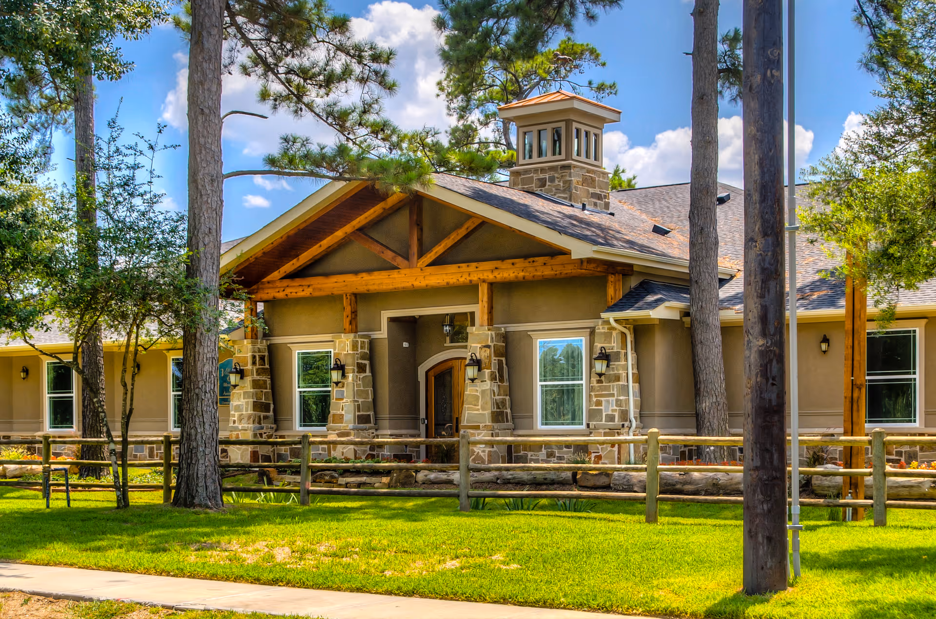Front exterior of a single-story stone-and-wood memory care building with a covered entrance, trees, and a manicured lawn.