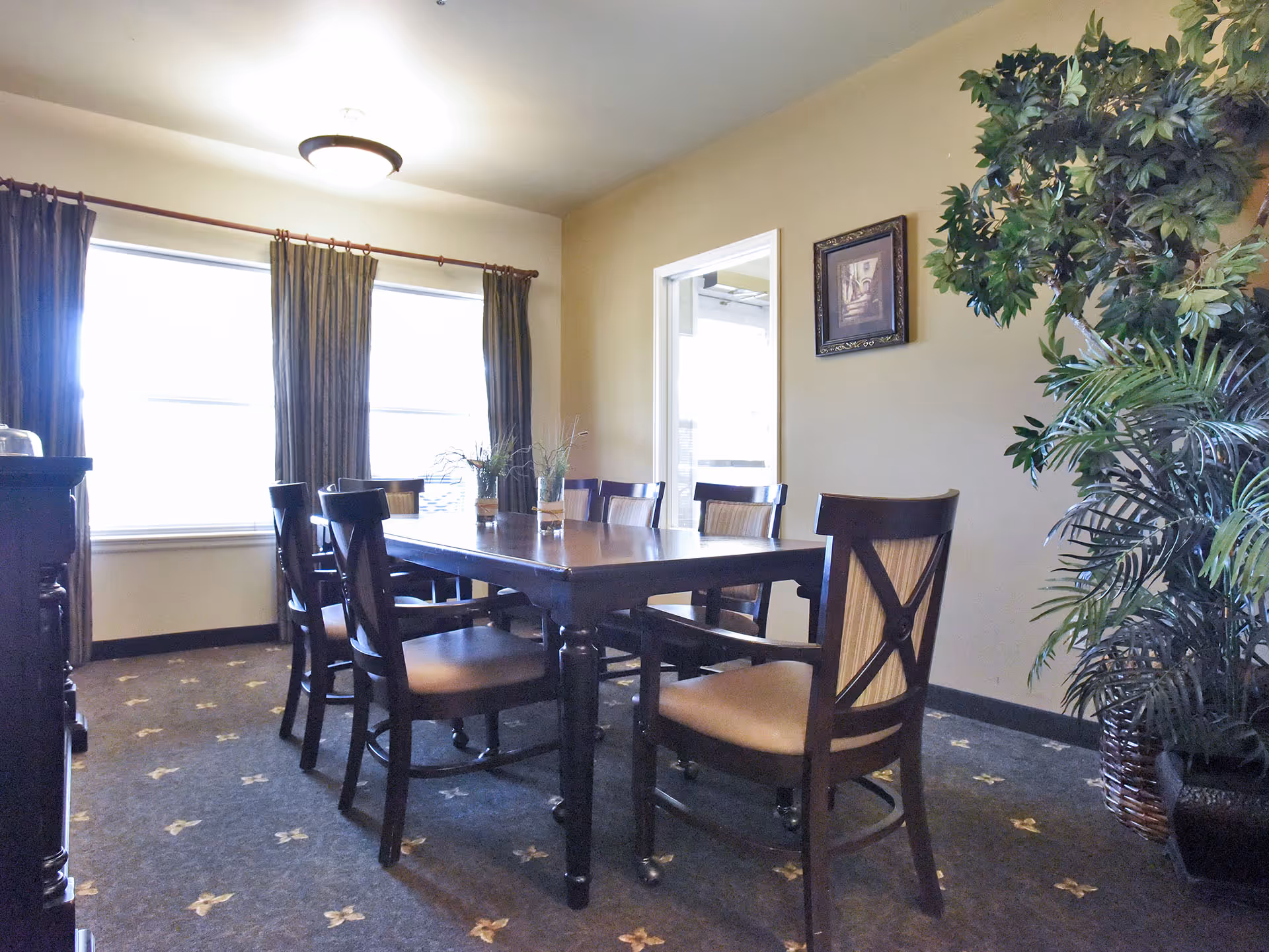 Dining room with a wooden table and chairs, large curtained windows, and potted plants.