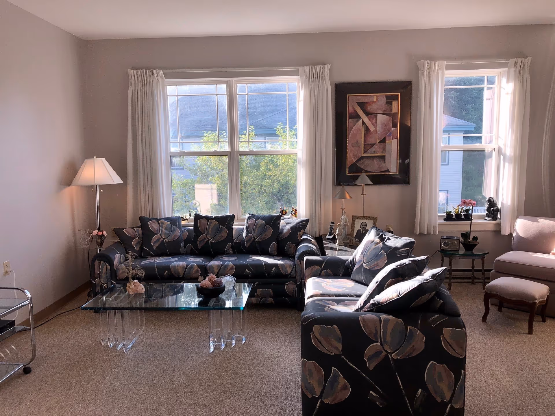 A cozy living room with two floral-patterned sofas facing each other, a glass coffee table in the center, a floor lamp on the left, and two large windows with white curtains letting in natural light. There is a framed abstract painting on the wall between the windows, a small side table with decorative items, and a pink armchair with an ottoman on the right.