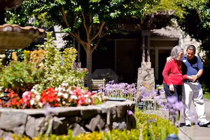 An elderly woman in a red sweater is walking outdoors with the assistance of a caregiver in a blue shirt and white pants. They are surrounded by a garden with colorful flowers, greenery, and a stone planter. Trees and a building structure are visible in the background.