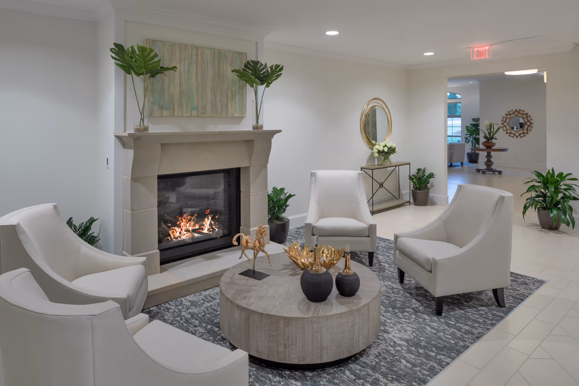 A cozy seating area with four white armchairs arranged around a round wooden coffee table with decorative items. There is a lit fireplace with a mantel decorated with two potted plants and a painting above it. The room has light-colored walls and flooring, with additional plants and a console table with a round mirror in the background.