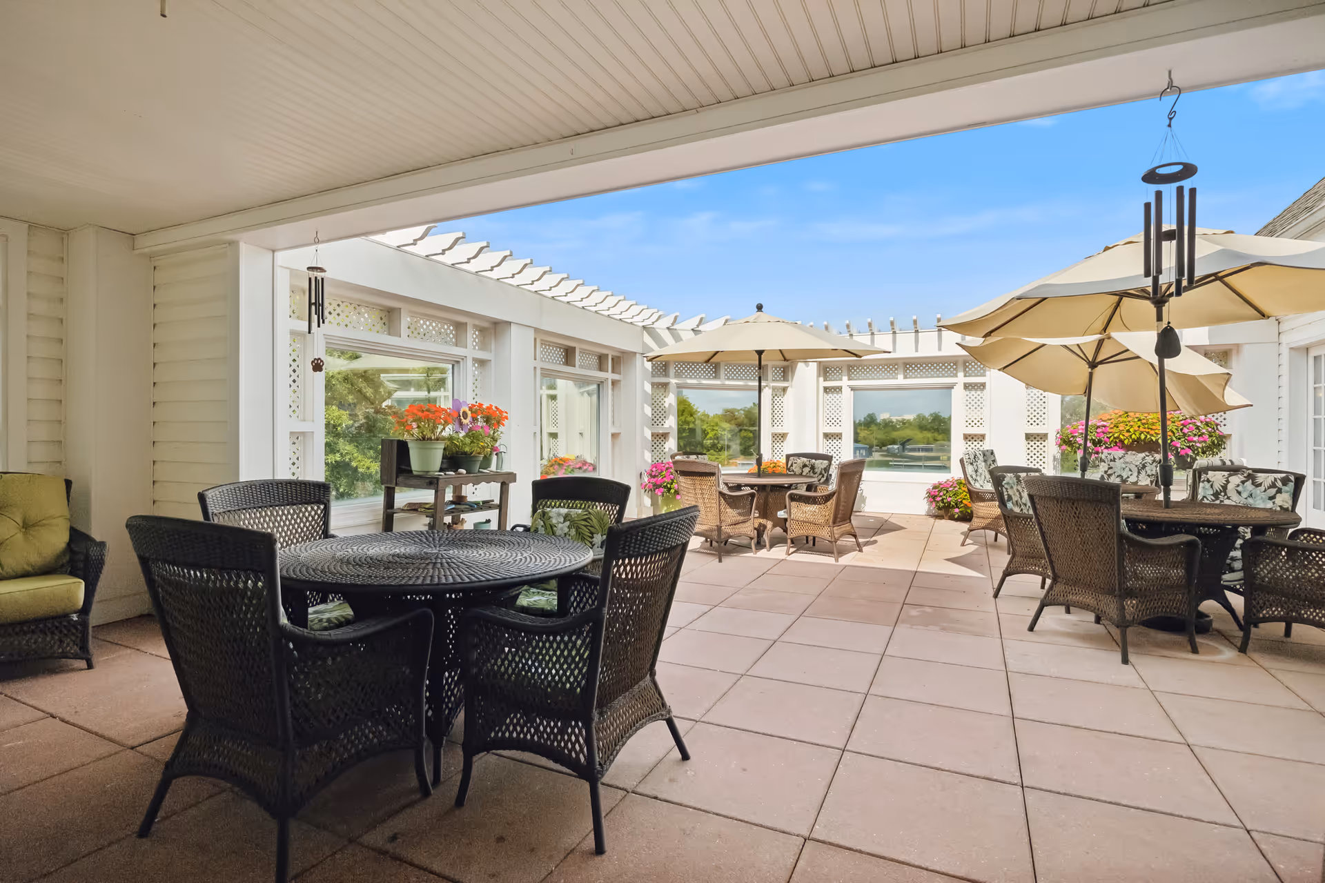 Outdoor patio area with multiple round tables and wicker chairs, some tables shaded by large beige umbrellas. There are potted flowers on a small table and around the patio, with a clear blue sky visible in the background.
