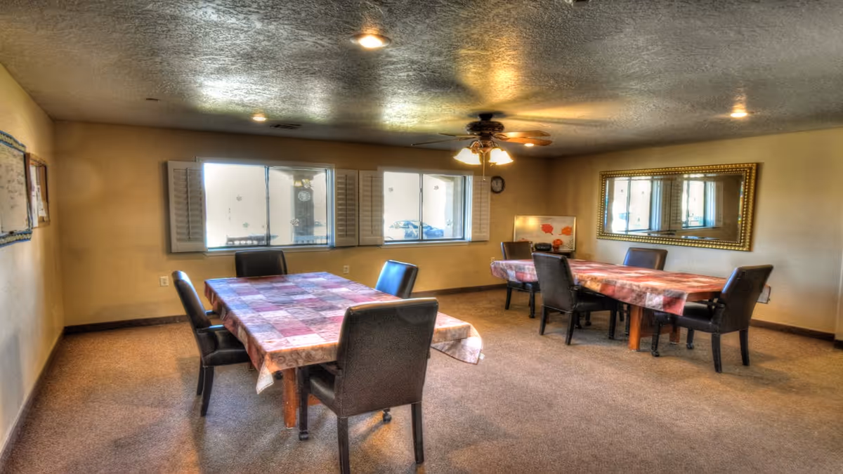 A room with two tables covered with patterned tablecloths, each surrounded by black chairs. The room has beige walls, carpeted floor, a ceiling fan with lights, a large mirror on one wall, and windows with white shutters letting in natural light.