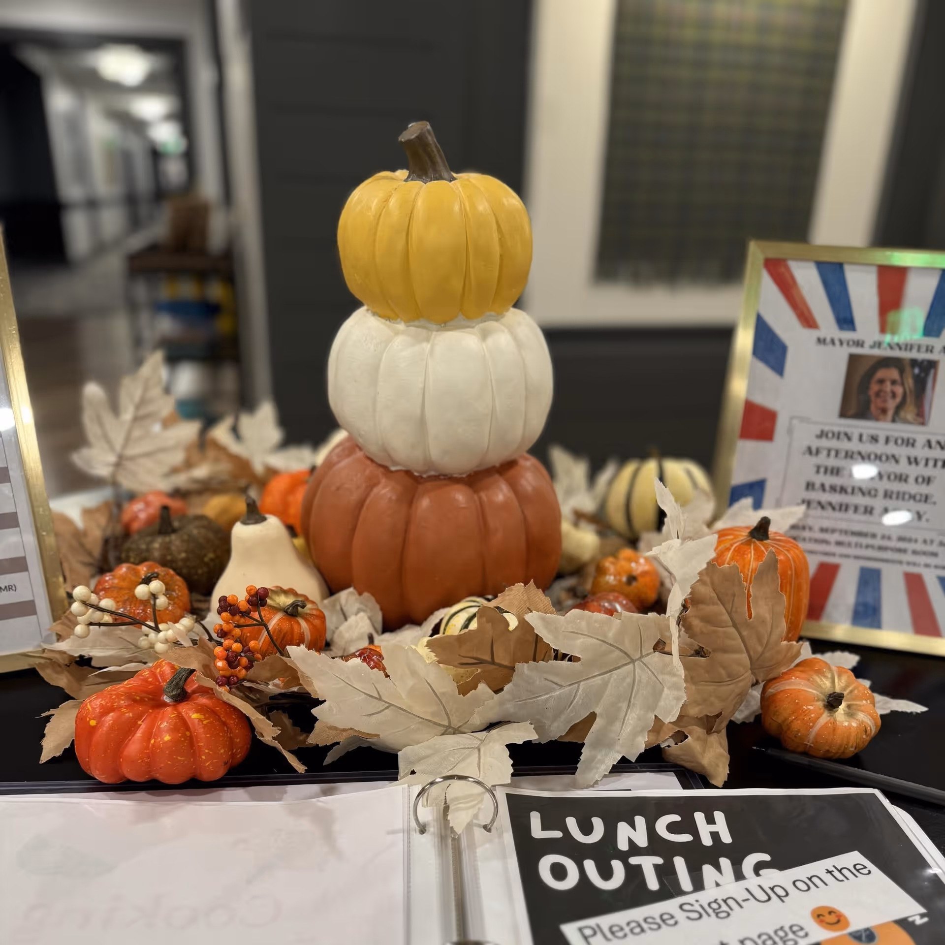 A fall-themed table centerpiece featuring a stack of three decorative pumpkins in yellow, white, and orange, surrounded by smaller pumpkins, autumn leaves, and berries. In the background, there is a framed sign and a binder with a 'Lunch Outing' sign-up sheet.