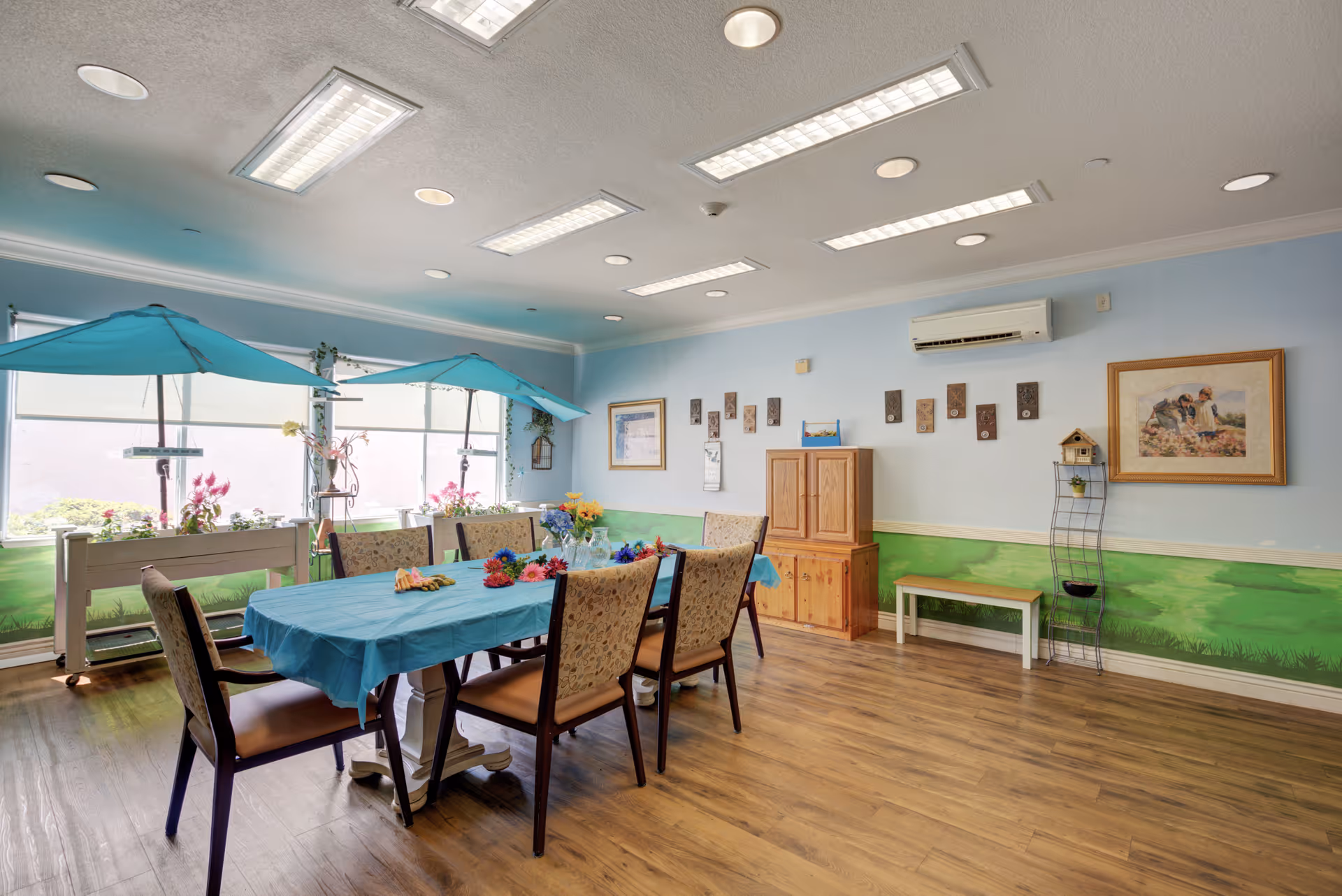 A bright and cheerful dining room with a long table covered by a blue tablecloth surrounded by eight cushioned chairs. The room has light blue walls with a green mural along the bottom half, wooden flooring, and large windows with blue umbrellas and flower boxes outside. The walls are decorated with framed pictures and small art pieces, and there is a wooden cabinet and a small bench against one wall.