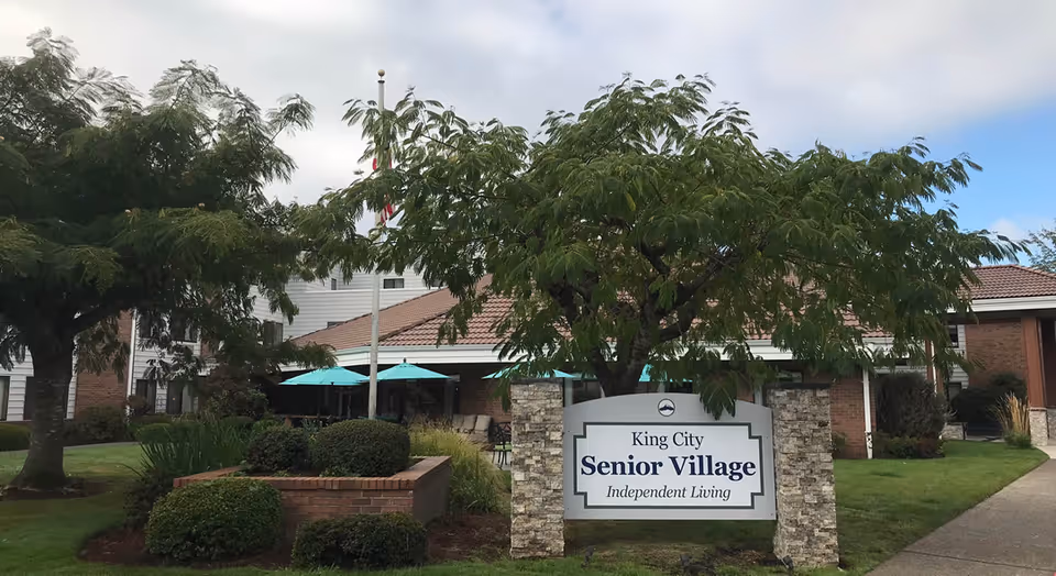 Outdoor view of King City Senior Village showing a building with a tiled roof, surrounded by green trees and bushes. There is a sign in front that reads 'King City Senior Village Independent Living'. Blue umbrellas are visible in the background near the building.