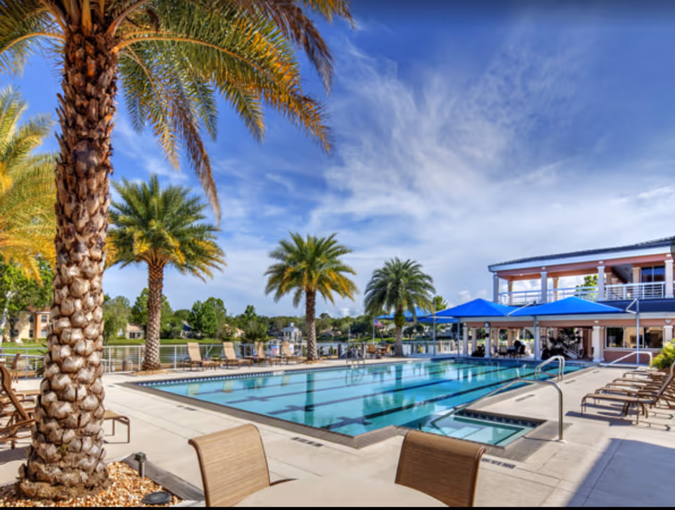 Outdoor swimming pool area with lounge chairs and palm trees under a blue sky with some clouds. A two-story building with blue awnings is visible in the background.