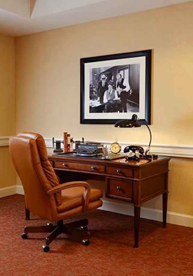 A cozy office corner with a brown leather swivel chair and a wooden desk. On the desk are a vintage typewriter, an old-fashioned telephone, a desk lamp, a clock, and some books. A black and white framed photograph of three men is hung on the beige wall above the desk.