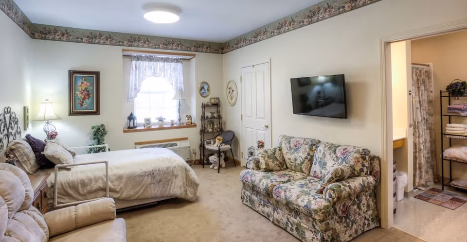 A cozy bedroom in a senior living facility featuring a bed with floral bedding, a floral-patterned couch, a recliner chair, a wall-mounted TV, a window with lace curtains, and a small shelving unit with decorative items. There is an open doorway leading to a bathroom with towels on a rack and a shower curtain.