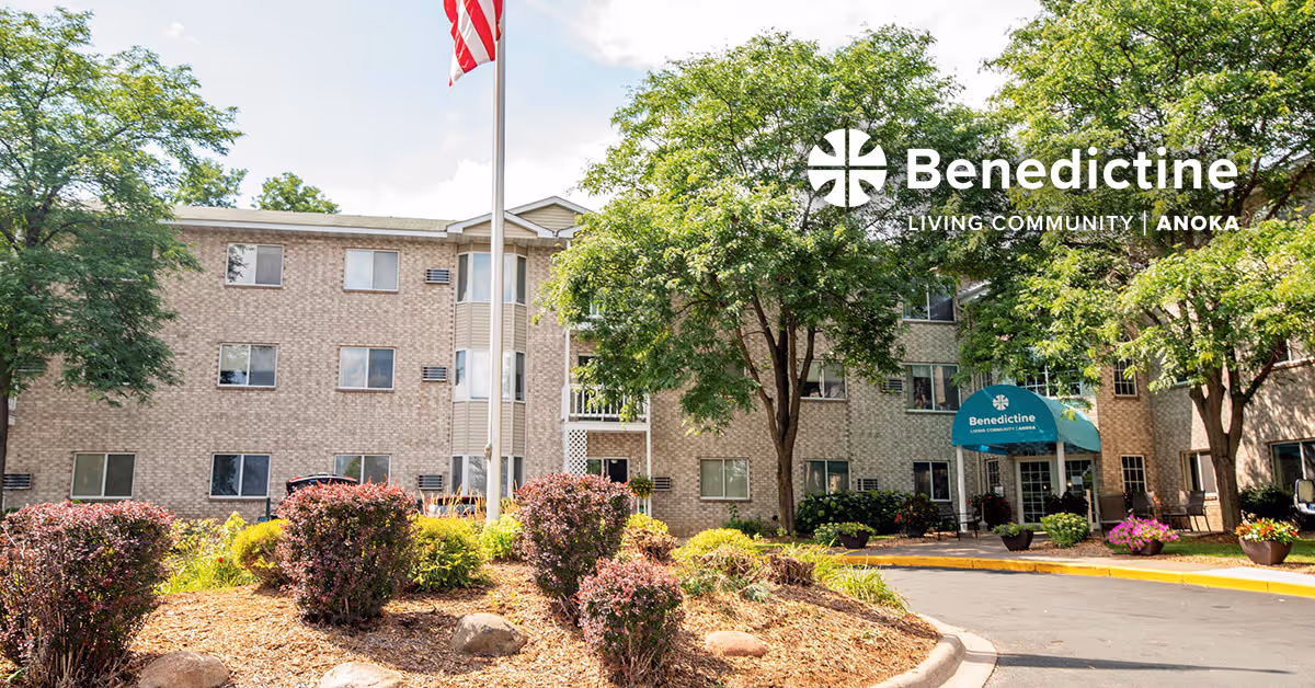 Exterior view of Benedictine Living Community-Anoka, a three-story brick building surrounded by trees and landscaped bushes, with an American flag on a flagpole in front and a blue awning over the entrance.