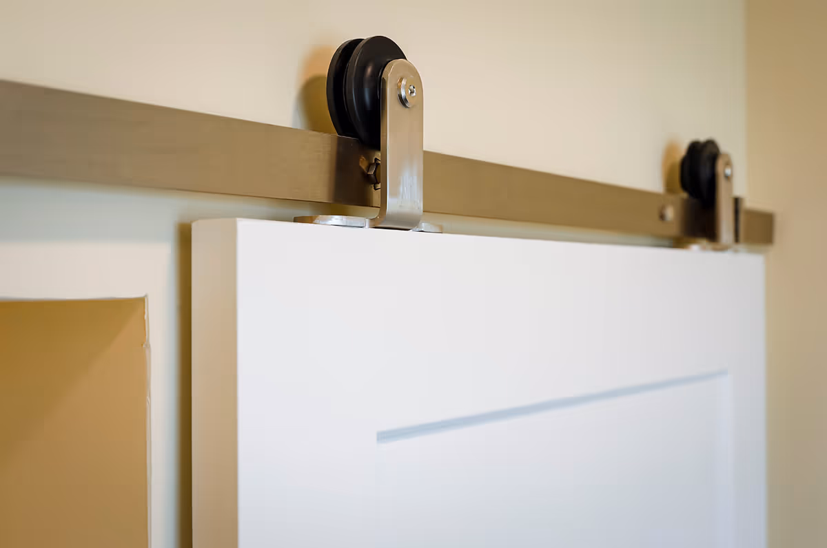 Close-up view of a sliding barn door hardware with metal rollers mounted on a beige wall, holding a white door panel.