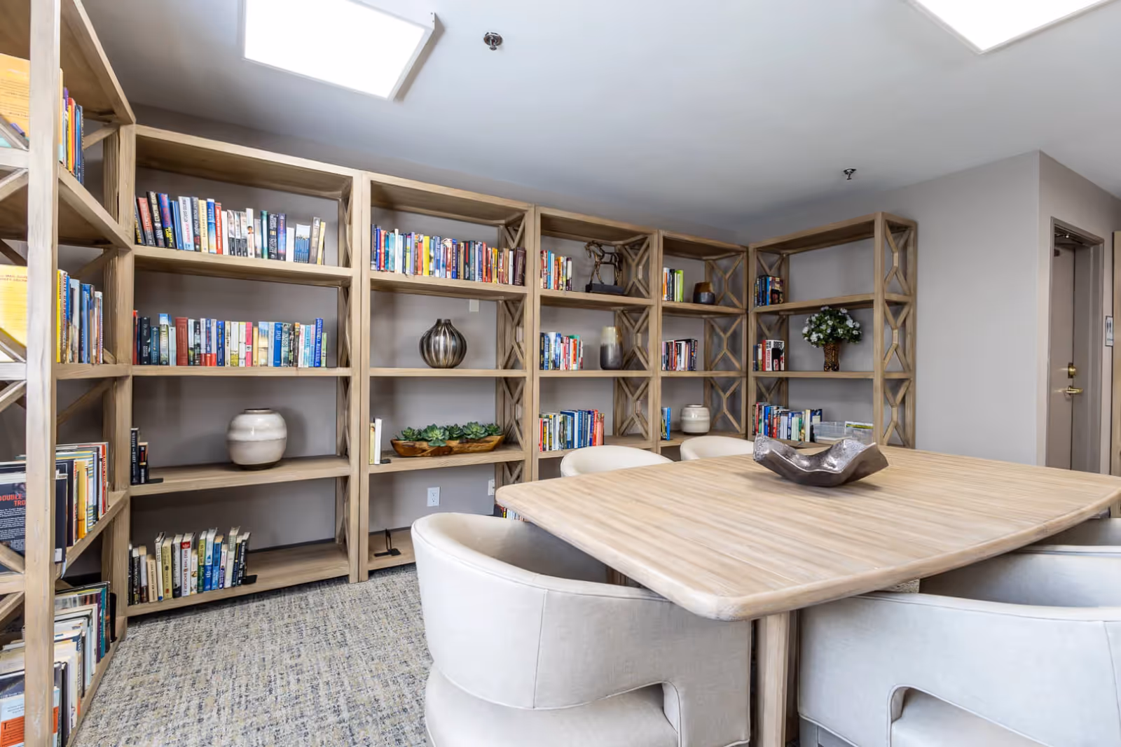 A cozy reading room with light wooden bookshelves filled with books and decorative items. In the center, there is a light wooden table surrounded by cushioned beige chairs. The room has a carpeted floor and neutral-colored walls with a door visible on the right side.