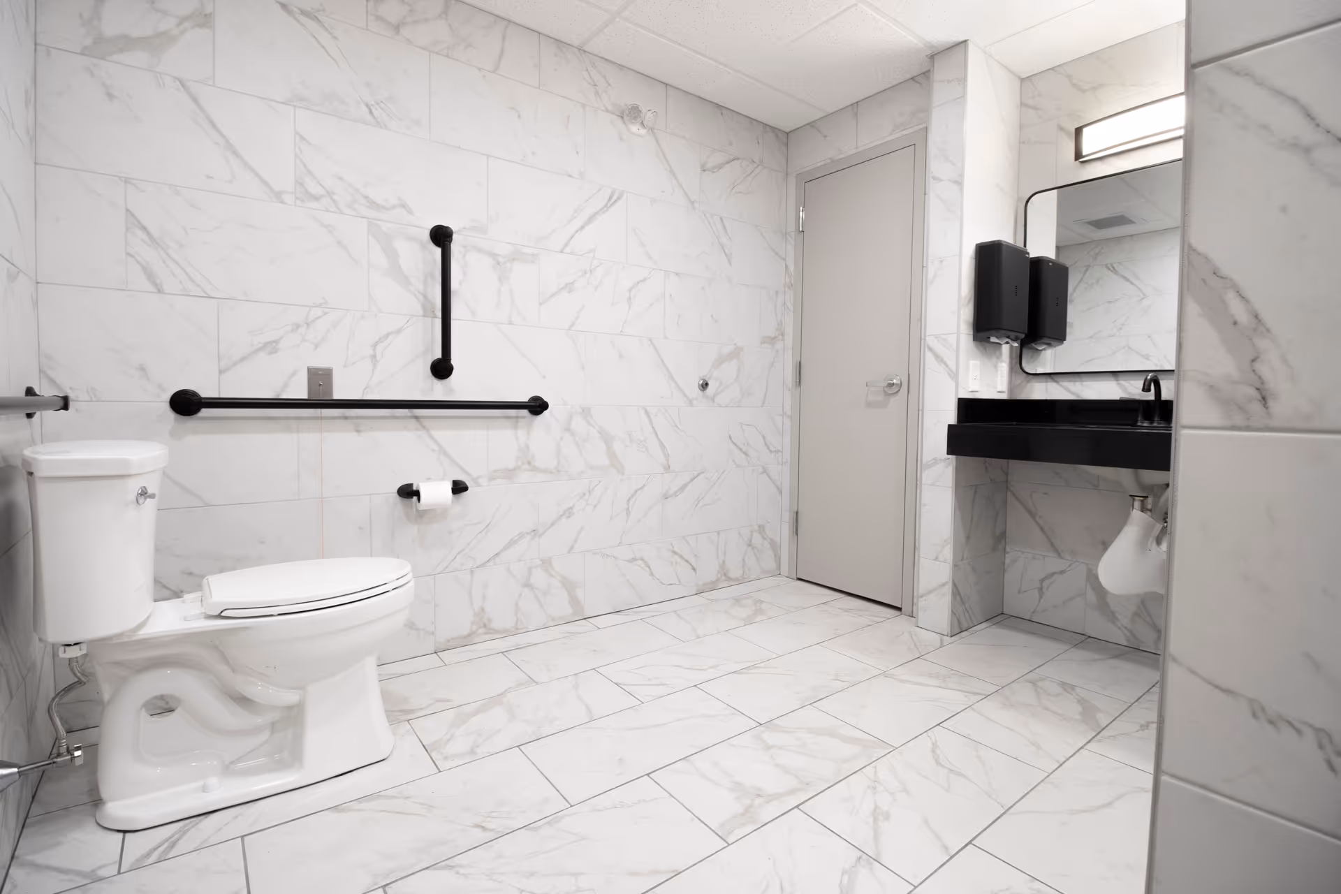 Accessible restroom with white marble tile, a toilet with grab bars, and a sink with mirror and soap dispensers.