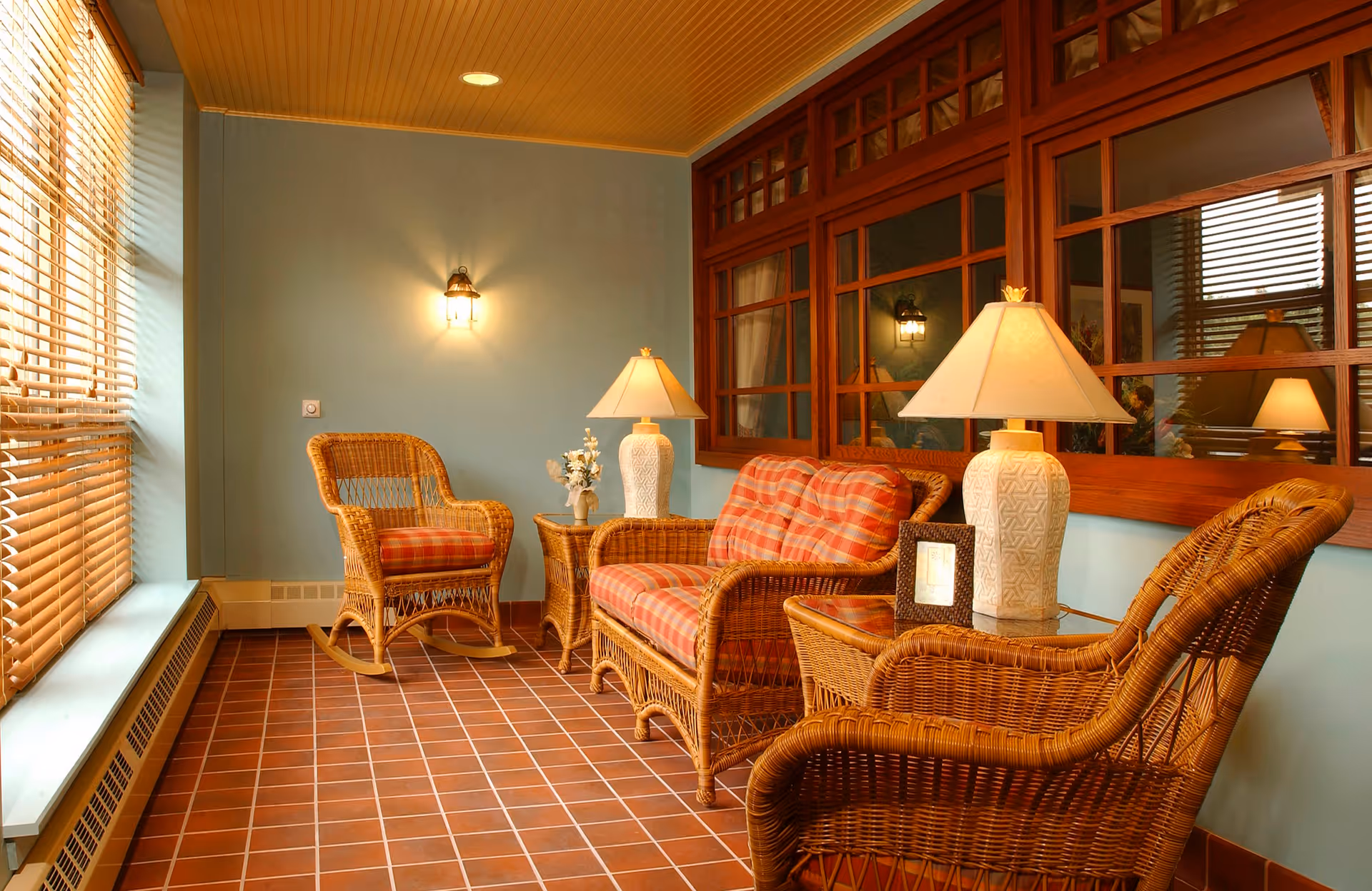 A cozy sitting area with wicker furniture including two armchairs and a loveseat with red plaid cushions, two side tables with white lamps, a small flower vase, and a framed photo. The room has a tiled floor, light blue walls, a wooden window with multiple panes, and a large window with wooden blinds letting in natural light.