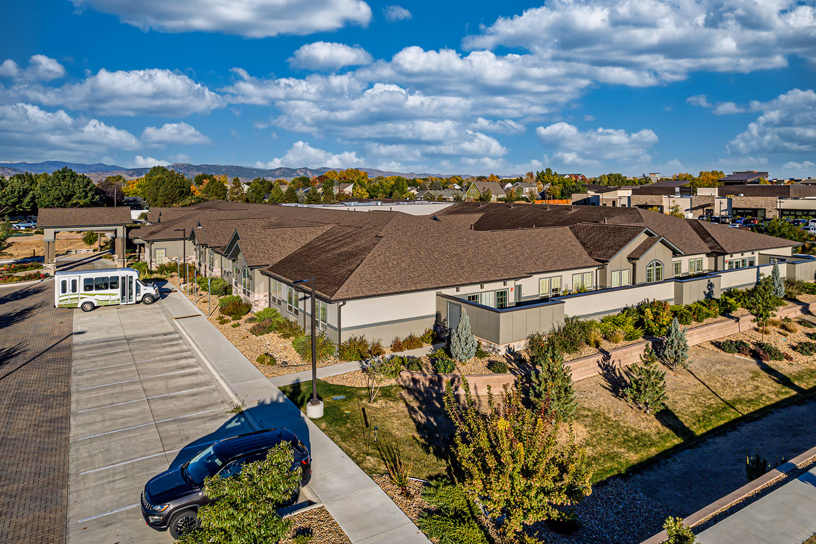 Aerial view of a single-story memory care facility building with a parking lot, shuttle bus, and landscaped grounds under a partly cloudy sky.