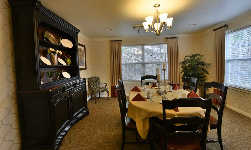 A dining room with a round table covered with a yellow tablecloth, set with glasses, cups, and red folded napkins. Six dark wooden chairs surround the table. A large dark wooden cabinet with decorative plates and glassware is against one wall. Two windows with beige curtains allow natural light into the room. A chandelier with multiple lights hangs from the ceiling, and a potted plant is in the corner.