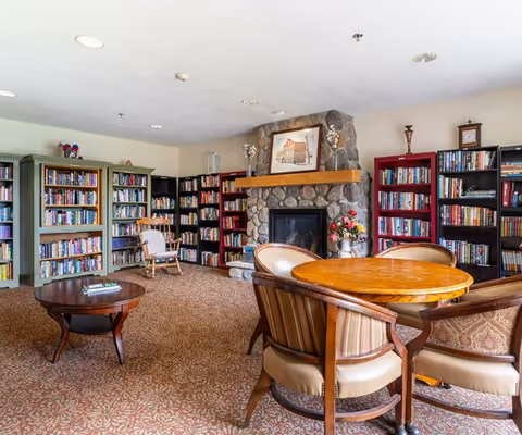 A cozy library room with multiple bookshelves filled with books along the walls, a stone fireplace with a wooden mantle and framed artwork above it, a round wooden table surrounded by four cushioned chairs, a wooden rocking chair, and a small round coffee table on a patterned carpeted floor.