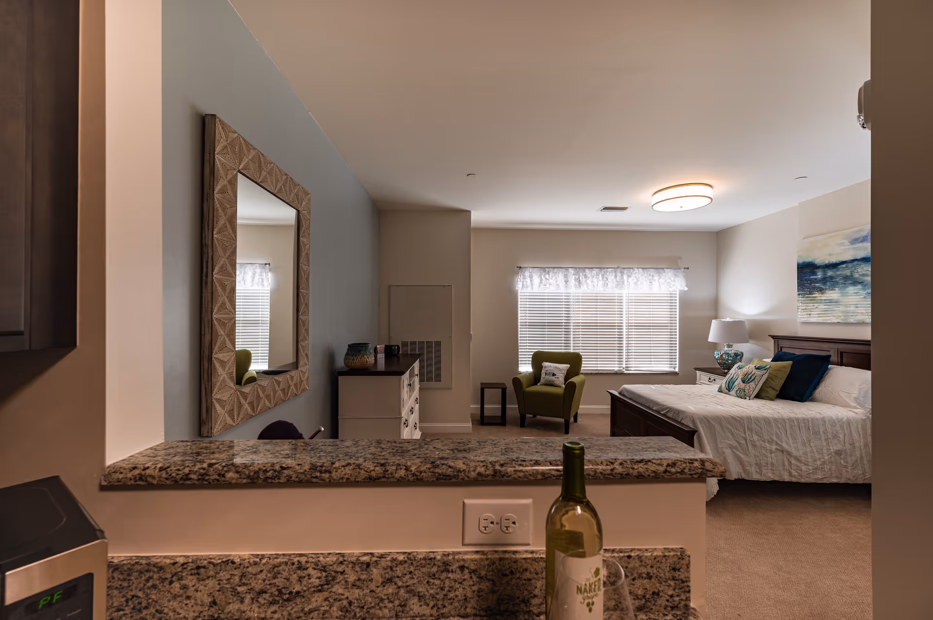 View from a kitchen counter into a bedroom area with a bed, green armchair, dresser, and a large window with blinds and a sheer curtain. The room has neutral-colored walls, a ceiling light fixture, and decorative pillows on the bed. A bottle of wine and a glass are on the granite kitchen counter in the foreground.