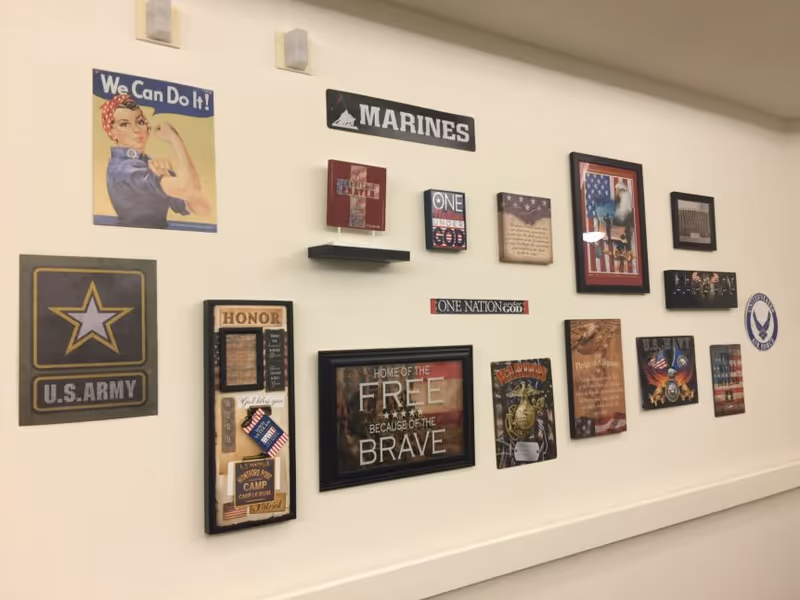 A wall decorated with various military-themed posters and plaques including a 'We Can Do It!' Rosie the Riveter poster, U.S. Army and Marines signs, patriotic quotes, and American flag imagery.