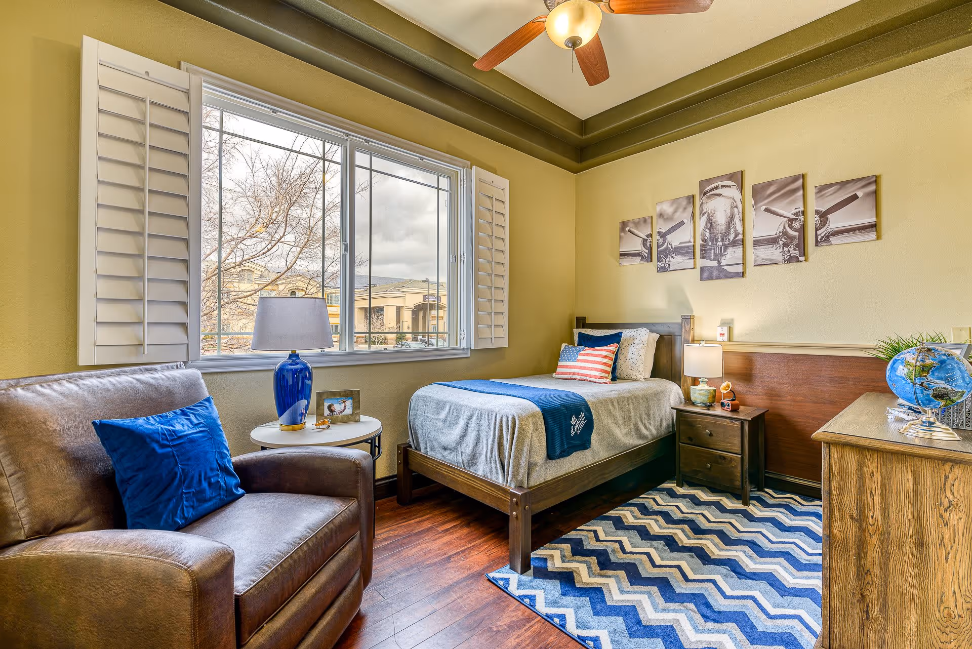A cozy bedroom in a senior living facility with a single bed dressed in gray and blue bedding, an armchair with a blue pillow, a small round side table with a blue lamp and a photo frame, a wooden nightstand with a lamp and small decorative items, a wooden dresser with a globe and plant, a large window with white shutters, and a ceiling fan with a light. The walls are painted light yellow and decorated with a five-panel airplane-themed artwork. The floor has a blue and white zigzag patterned rug.