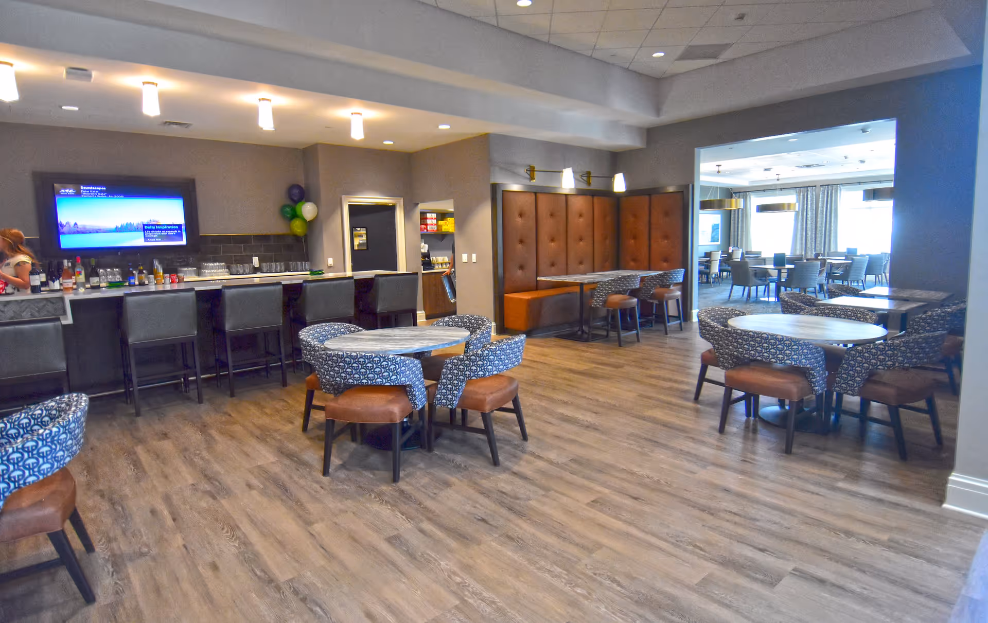Interior view of a senior living community dining and lounge area with multiple round tables surrounded by patterned chairs, a bar with high stools, a wall-mounted TV, and a booth seating area with brown cushioned panels. The space has wood flooring and modern lighting fixtures.