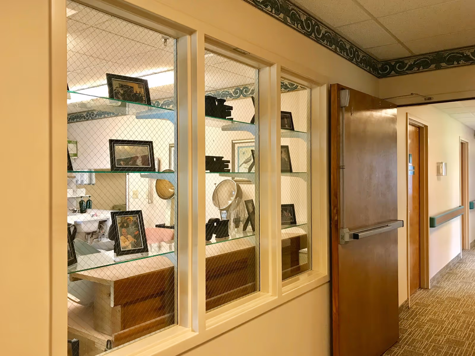 View through a glass partition with wire mesh showing a room with framed photos on glass shelves and a sink in the background, adjacent to a hallway with carpeted floor, wooden doors, and a handrail along the wall.