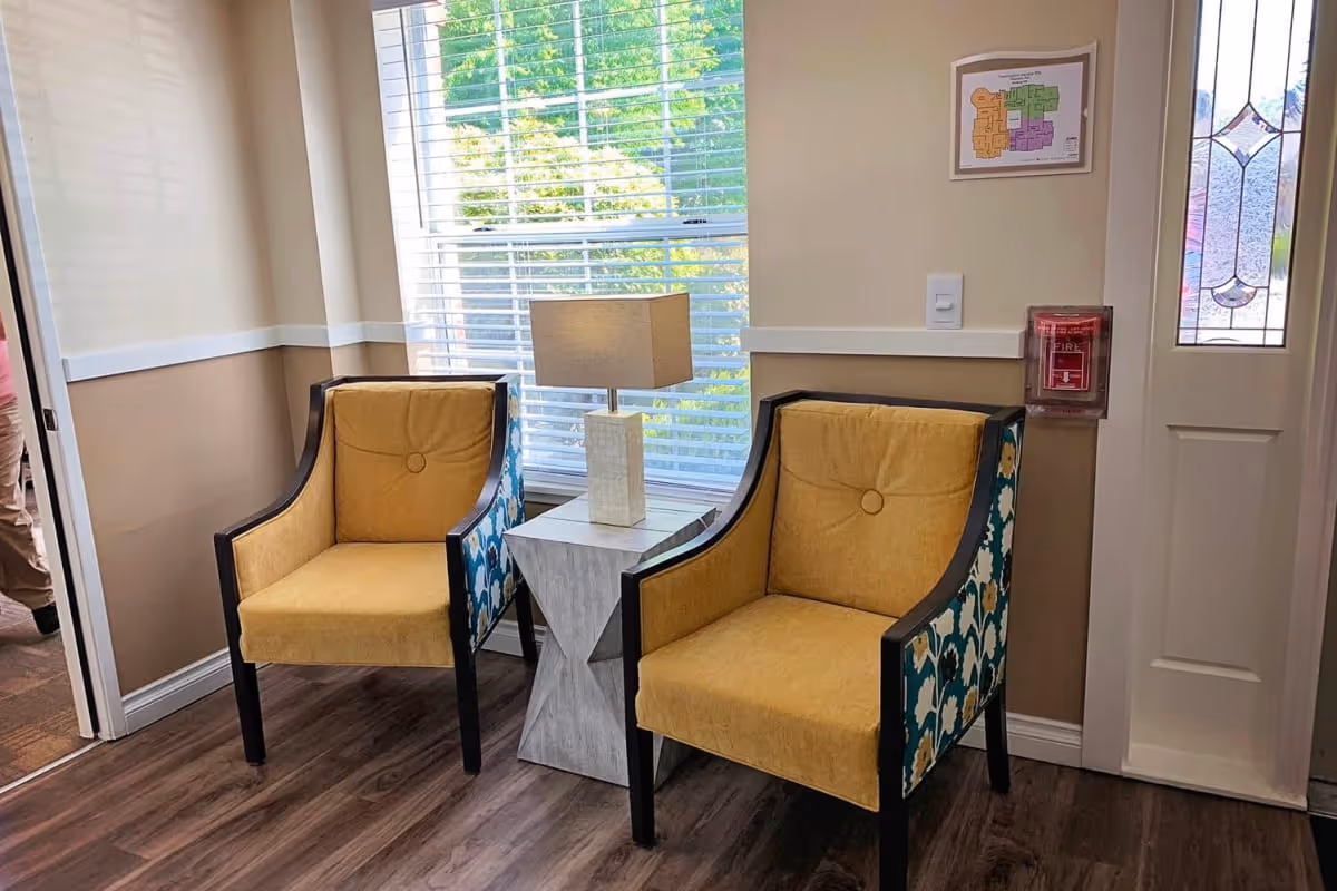 Two yellow upholstered chairs with patterned sides flank a small table and lamp in a sunny seating area by a window and entry door.