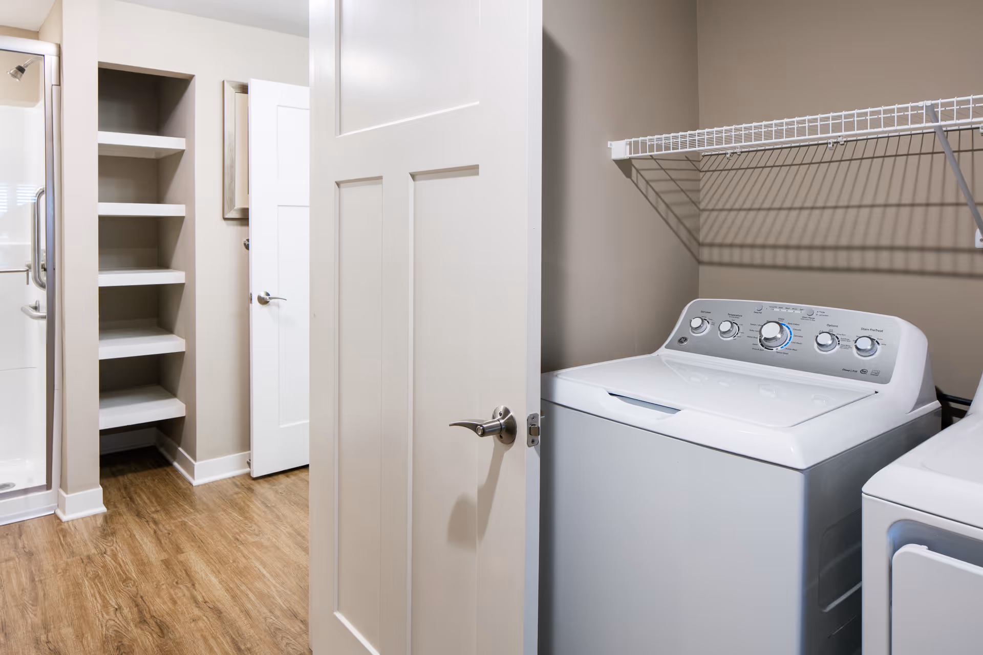 Laundry room with a white washing machine and dryer, a wire shelf above them, wooden floor, and an open door leading to a hallway with built-in shelves and a shower area.