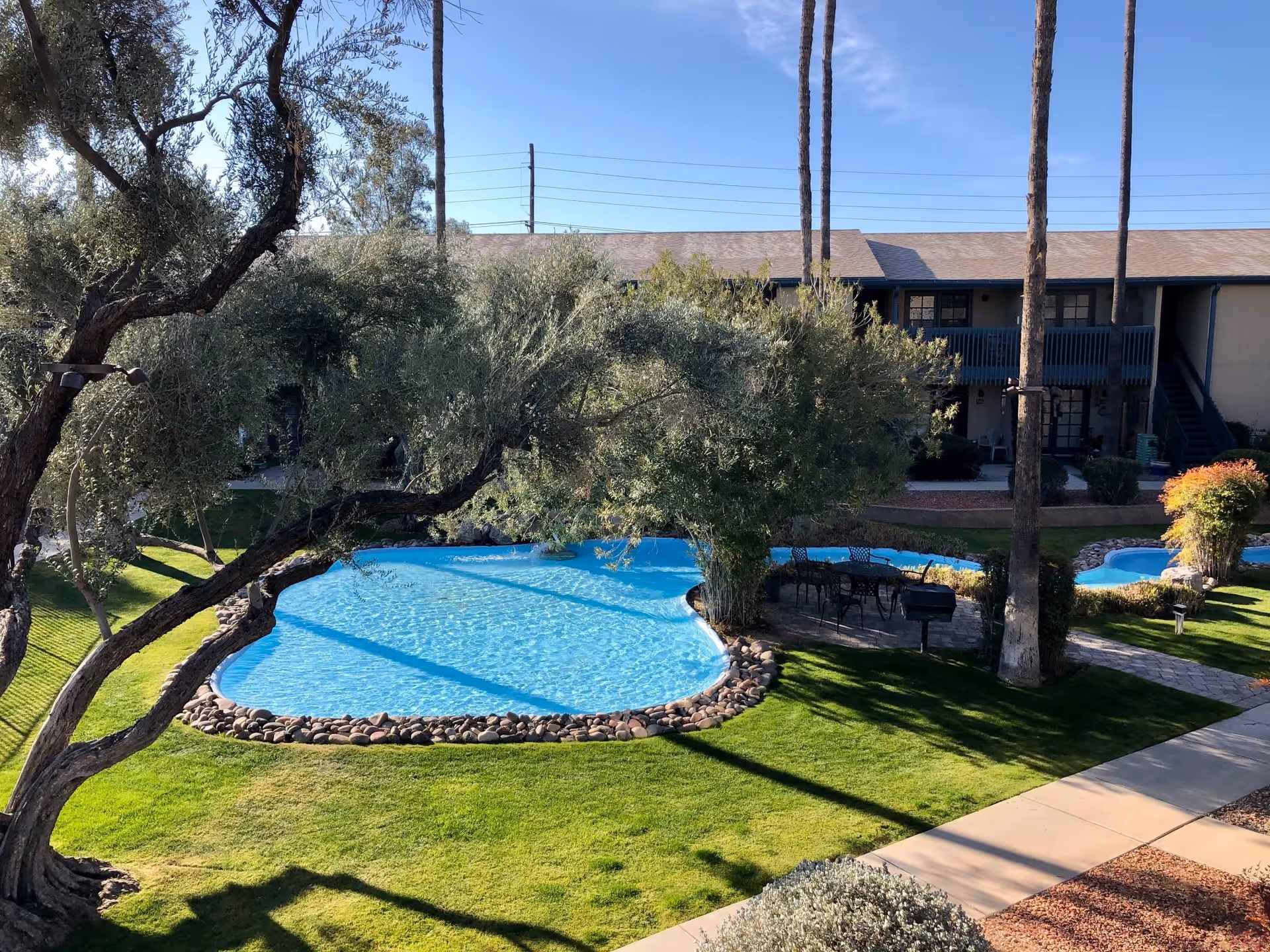 Outdoor view of a residential facility with a kidney-shaped swimming pool surrounded by green grass, trees, and a paved walkway. There is a seating area with a table and chairs near the pool, and a two-story building with balconies and stairs in the background under a clear blue sky.
