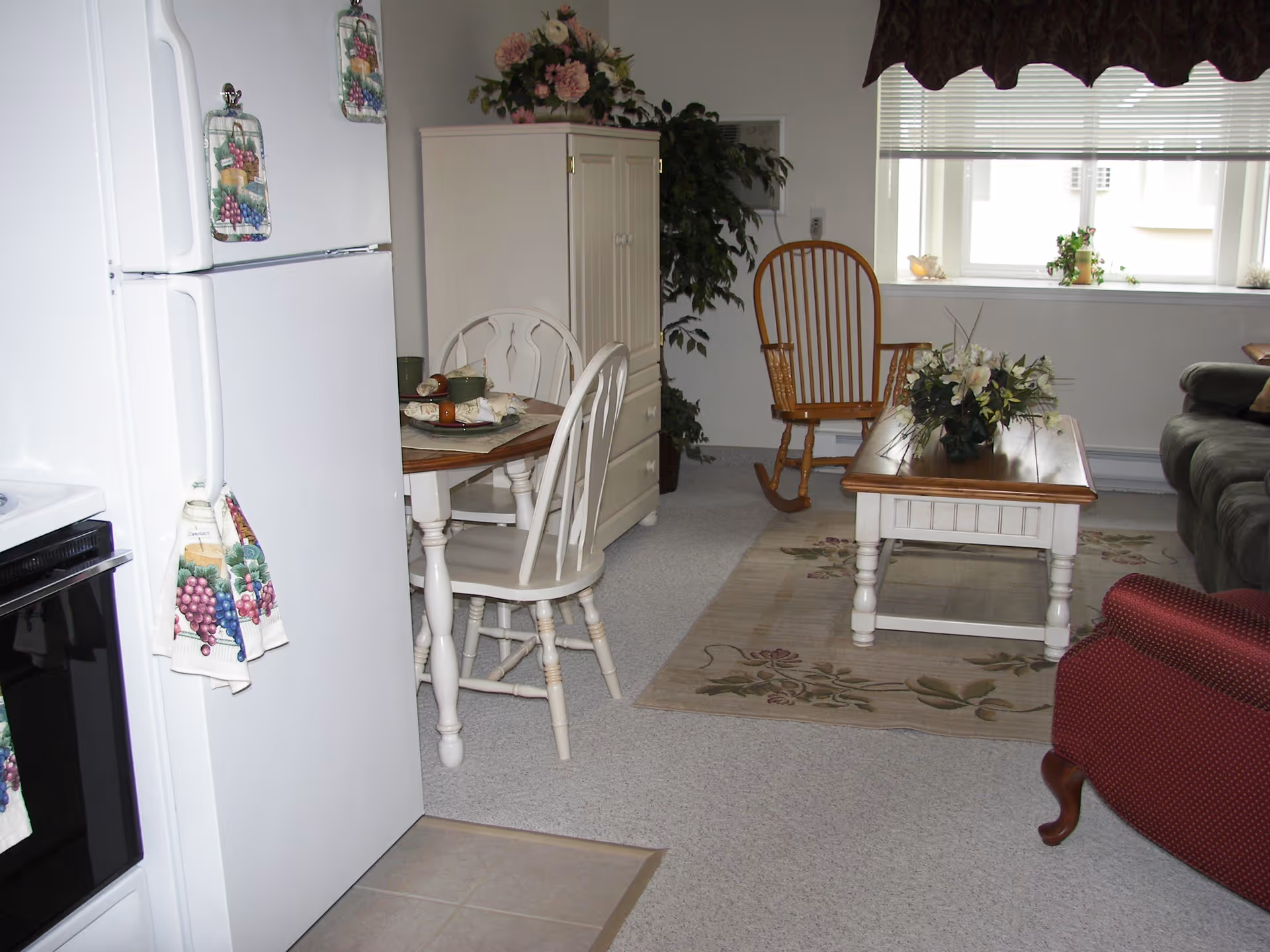 A cozy living space featuring a white refrigerator and stove on the left, a small wooden dining table with two chairs, a wooden rocking chair near a window with blinds, a coffee table with a floral arrangement, a green sofa, and a red armchair. The room has light-colored carpet and a floral area rug under the coffee table. There are decorative items including flowers on a cabinet and plants near the window.