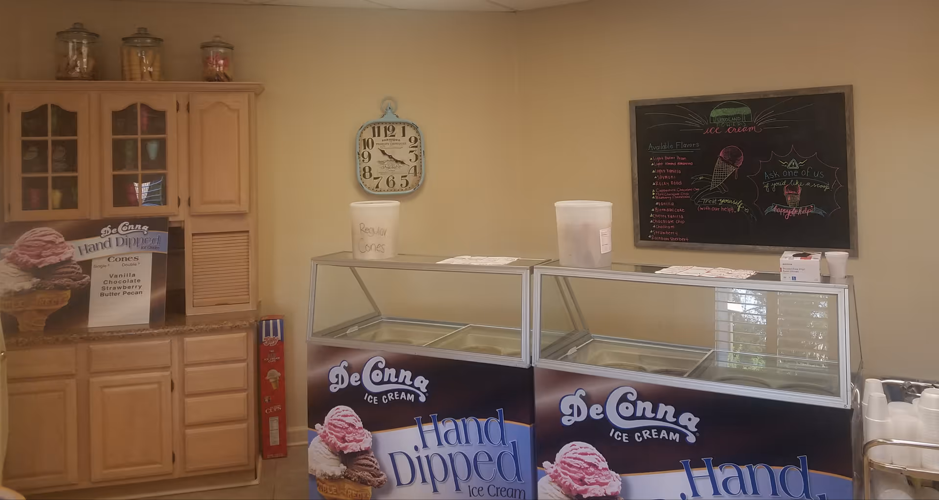 Ice cream serving area with two DeConna Hand Dipped Ice Cream freezers, a wooden cabinet with glass doors containing cups and cones, a wall clock, and a chalkboard menu listing available ice cream flavors and a message inviting people to ask for help.
