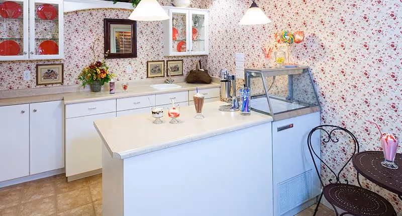 A cozy kitchen area with white cabinets and countertops, floral wallpaper, and a small ice cream or dessert counter. There are three dessert glasses with different treats on the counter, a vase with flowers, and a small round table with two metal chairs. The space is well-lit with two hanging pendant lights.