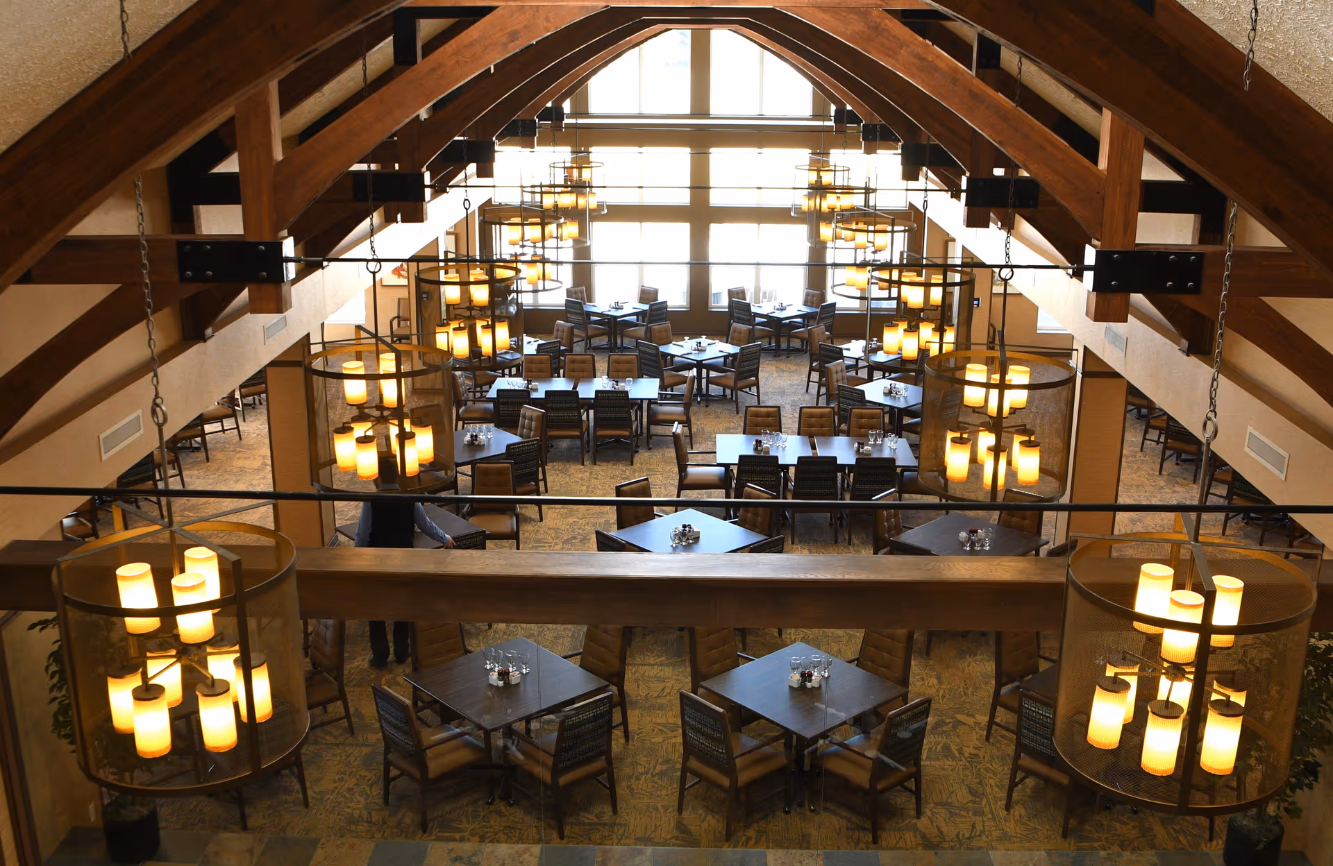 Interior view of a spacious dining room with multiple tables and chairs arranged neatly. The room features large windows letting in natural light and has wooden beams and multiple hanging chandeliers with warm lighting.