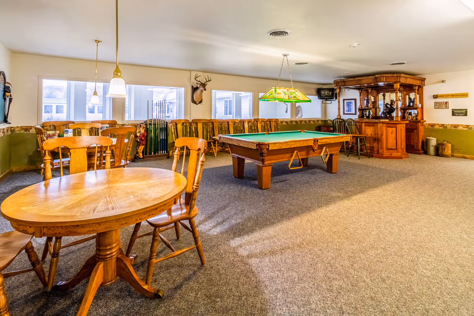 A recreational room with a pool table in the center, surrounded by wooden chairs and tables. There is a wooden bar in the back right corner, a mounted deer head on the wall, and several windows letting in natural light. The room has carpeted flooring and a green and beige color scheme on the walls.