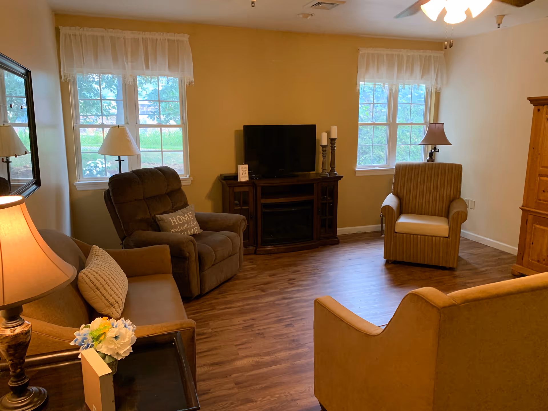 A cozy living room with wooden flooring, featuring a brown recliner chair with a pillow, a striped armchair, a beige sofa, a wooden TV stand with a flat-screen TV and decorative candles, two windows with sheer white curtains, and several lamps providing warm lighting.