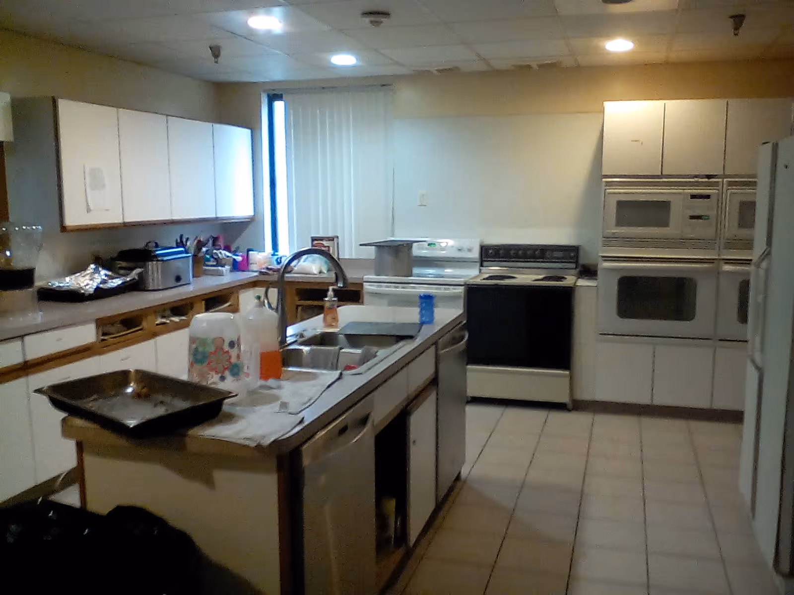 Interior view of a kitchen with white cabinets, a stove, oven, microwave, refrigerator, and a sink on a central island. Various kitchen items and cleaning supplies are on the countertops.