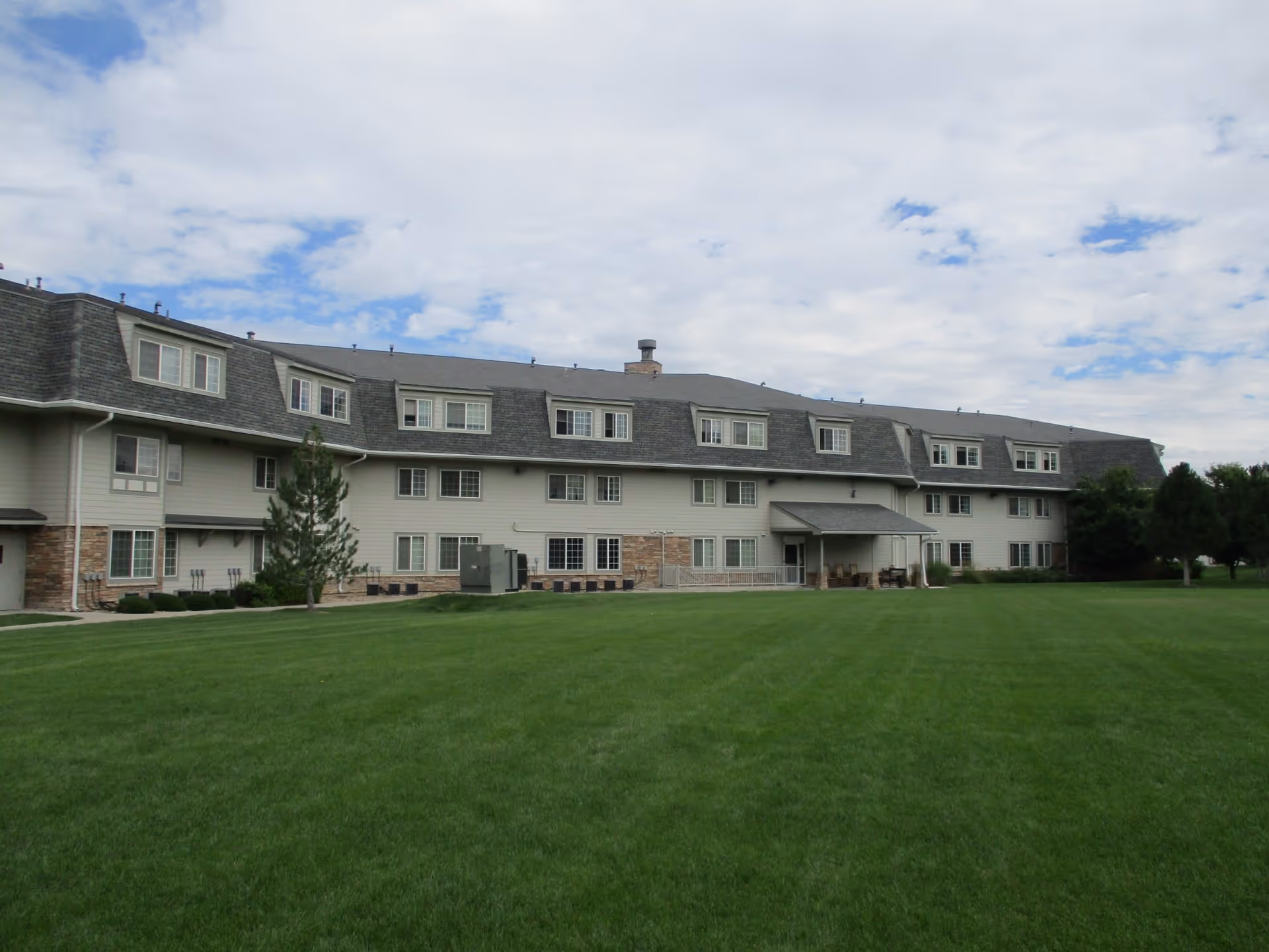 Exterior view of a large, multi-story senior living facility building with multiple windows and a covered entrance, surrounded by a well-maintained green lawn under a partly cloudy sky.