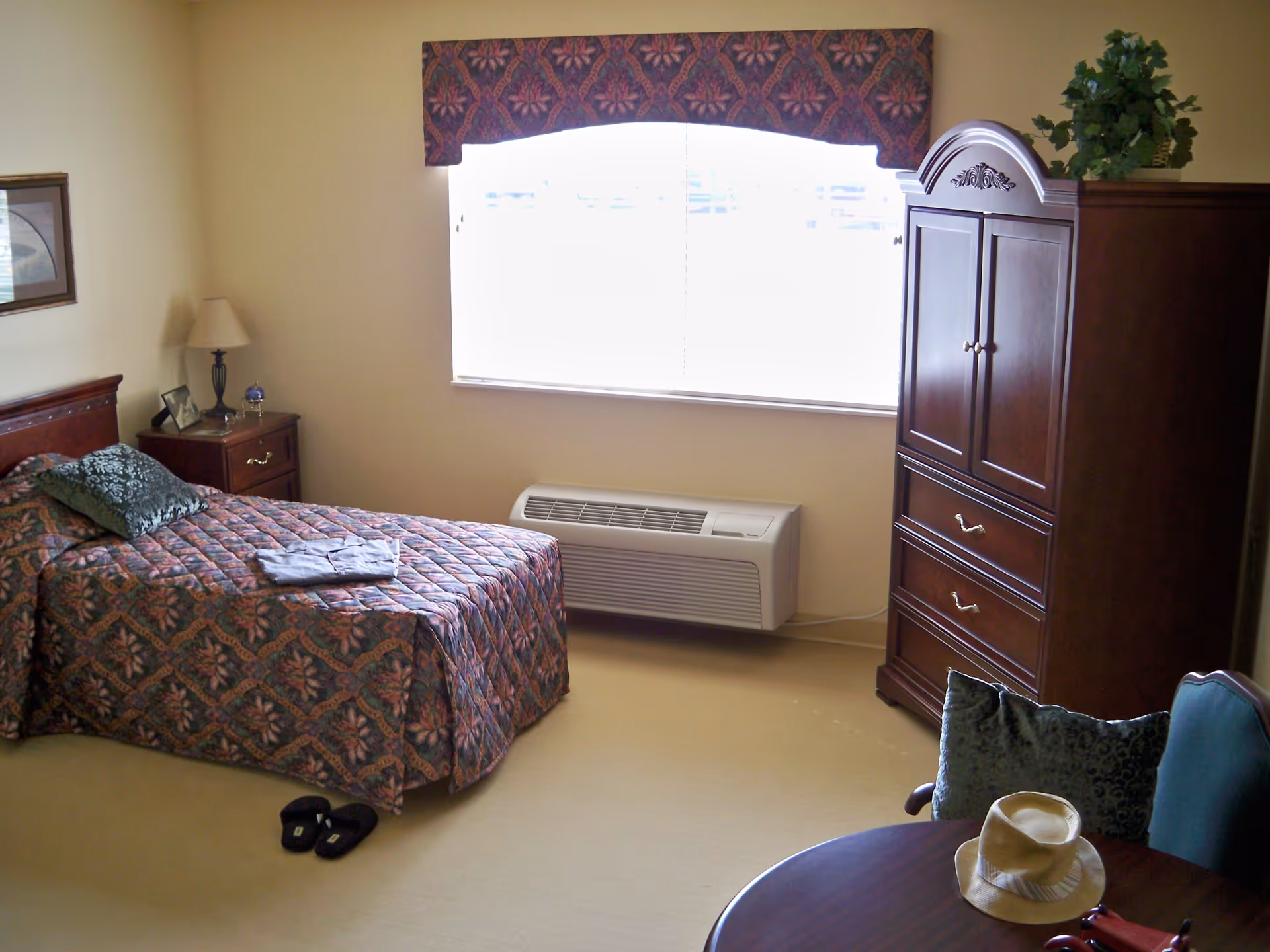 Furnished bedroom with a single bed, nightstand, armoire and window in a senior living facility.