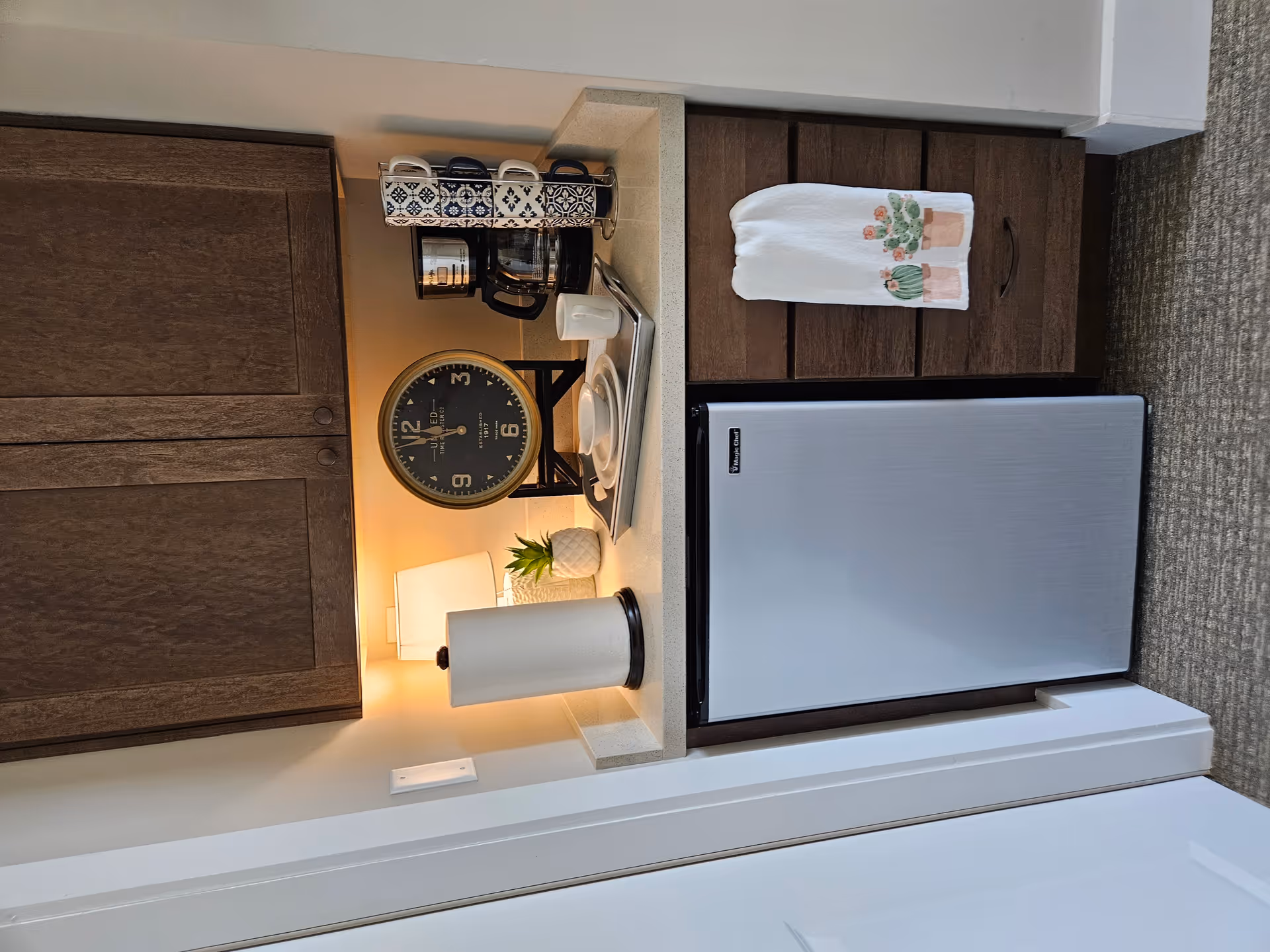 A small kitchenette area with a mini refrigerator, wooden cabinets, and a countertop. On the countertop, there is a paper towel holder, a small lamp, a decorative plant, a large round clock, a coffee maker, a set of stacked mugs on a stand, and a tray with dishes and a mug. A white towel with cactus designs hangs from a drawer handle.