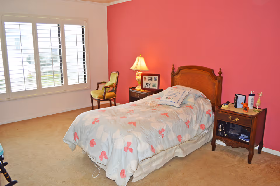 Sunlit bedroom with a single bed against a pink wall, wooden nightstands, a lamp, a chair, and a large window with shutters.