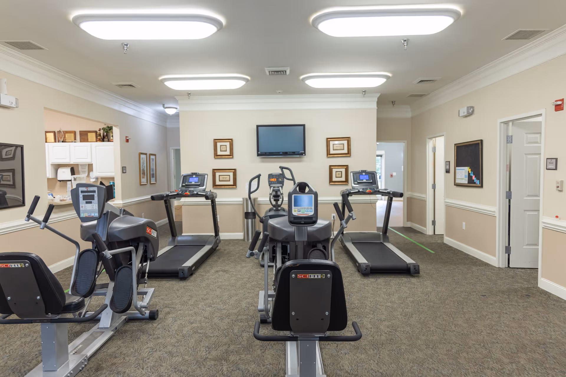 A fitness room in a senior living facility with exercise equipment including treadmills, a recumbent bike, and an elliptical machine. The room has beige walls, carpeted floor, framed pictures on the wall, and a flat-screen TV mounted on the wall.